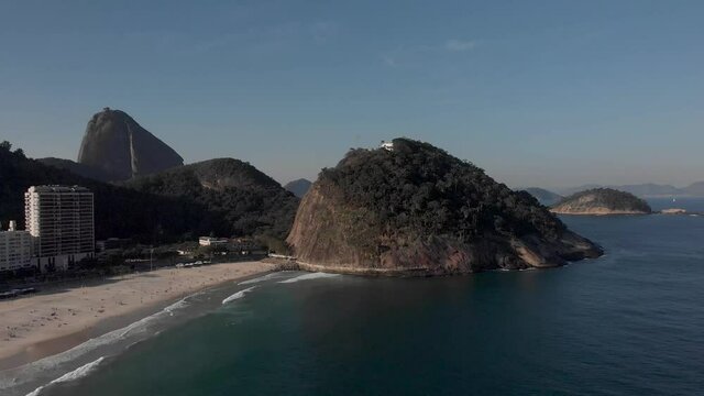 Steady aerial view of Leme beach at the end of Copacabana boulevard with people on the beach, the fort on top of the hill and Sugarloaf mountain in the background against a clear blue sky