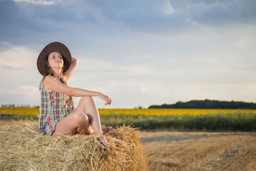Naklejka na ściany i meble Young girl in a cowboy hat on hay bale. Girl farmer in denim shorts and plaid shirt, in a wheat field in sunset. Happy Woman cowgirl on a farm. Freedom lifestyle.