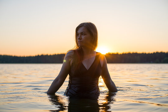 Beautiful Russian girl in a long black swimsuit swims outside the city on the lake in the rays of sunset or dawn. Splashing water on the background of the sun. Happy girl.
