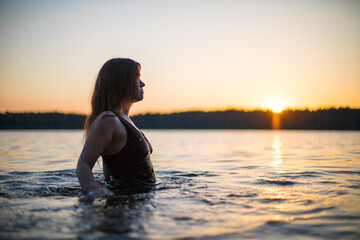 Beautiful Russian girl in a long black swimsuit swims outside the city on the lake in the rays of...