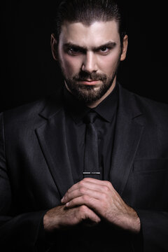 Young white Caucasian man on a black background in a black jacket, shirt and black tie looks out from under his forehead. Strict male portrait. The guy in the classic black suit.