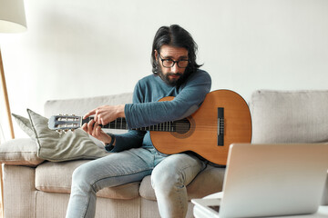 Young caucasian man sitting on sofa at home and adjusting acoustic guitar, watching guitar lesson online on laptop