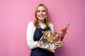 girl artist holds brushes and a palette and smiles on a pink background, student of art school,...