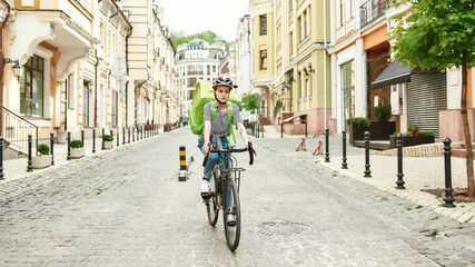 Young delivery woman in helmet with thermo bag or backpack riding a bike along the city, delivering...