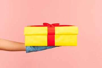 Profile side view closeup of human hand in blue surgical gloves holding yellow gift box. indoor,...