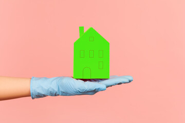 Fototapeta na wymiar Profile side view closeup of human hand in blue surgical gloves holding green paper house exterior in hand. indoor, studio shot, isolated on pink background.
