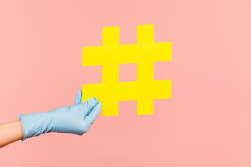 Profile side view closeup of human hand in blue surgical gloves holding yellow hashtag. indoor, studio shot, isolated on pink background.