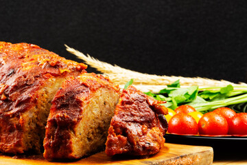 Classic meatloaf on a cutting board on a wooden background. In the background are red and yellow cherry tomatoes, parsley, ears of wheat
