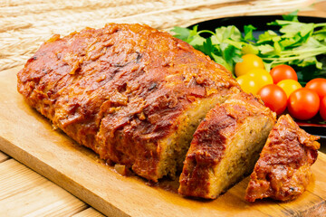 Classic meatloaf on a cutting board on a wooden background. In the background are red and yellow cherry tomatoes, parsley, ears of wheat