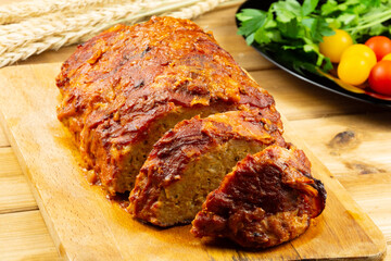 Classic meatloaf on a cutting board on a wooden background. In the background are red and yellow cherry tomatoes, parsley, ears of wheat