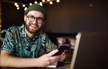Happy male hipster in earphones using laptop and smartphone looking at the camera and smiling