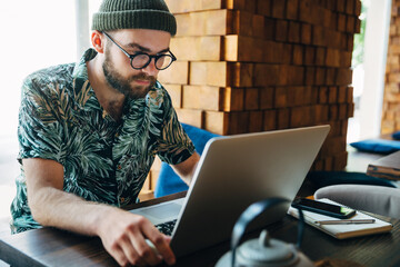 Bearded hipster businessman dressed in shirt wearing hat and eyeglasses working on laptop in cafe