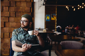 Modern fashionable male student with beard, hat and eyeglasses reading book drinking coffee