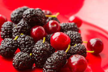 Ripe large black mulberries and red cherries on a red plate, close-up.