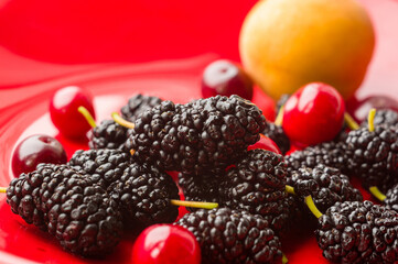 Ripe large black mulberries, peach and red cherries on a red plate, close-up.