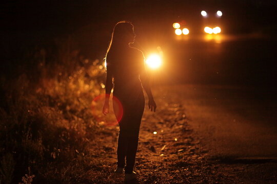 Silhouette of young slender woman in the backlight of car headlights on the road
