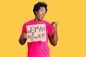 Handsome african american man with afro hair holding woman power banner pointing thumb up to the side smiling happy with open mouth