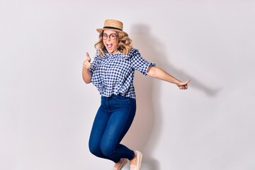 Young beautiful blonde woman wearing glasses and summer hat smiling happy. Jumping with smile on face over isolated white background.