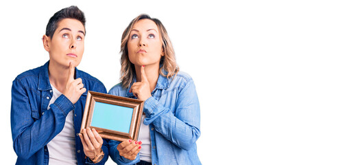 Couple of women holding empty frame thinking concentrated about doubt with finger on chin and looking up wondering