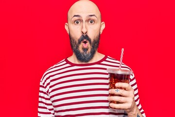 Young handsome man drinking glass of cola beverage scared and amazed with open mouth for surprise,...