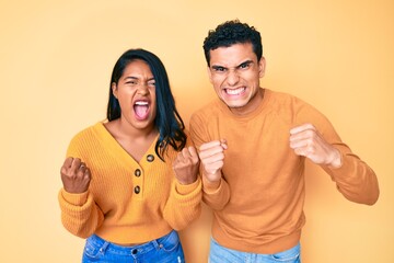 Beautiful latin young couple wearing casual clothes together angry and mad raising fists frustrated and furious while shouting with anger. rage and aggressive concept.