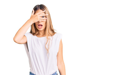 Beautiful caucasian woman with blonde hair wearing casual white tshirt peeking in shock covering face and eyes with hand, looking through fingers with embarrassed expression.