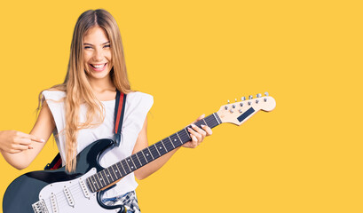 Fototapeta premium Beautiful caucasian woman with blonde hair playing electric guitar smiling happy pointing with hand and finger
