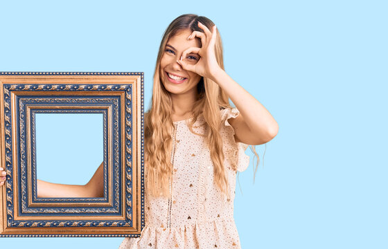 Beautiful caucasian woman with blonde hair holding empty frame smiling happy doing ok sign with hand on eye looking through fingers