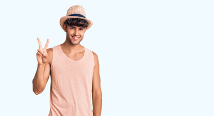 Young hispanic man wearing summer hat showing and pointing up with fingers number two while smiling...