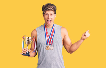 Young handsome man holding champion trophy wearing medals screaming proud, celebrating victory and success very excited with raised arms