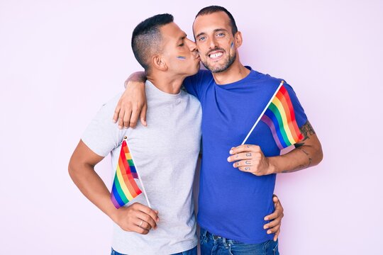 Young couple of gay men together in love holding homosexual pride colorful flag kissing with affection