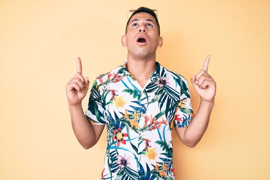 Young handsome latin man wearing summer clothes amazed and surprised looking up and pointing with fingers and raised arms.
