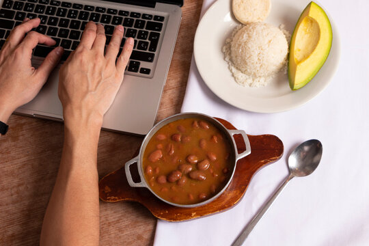 Frijoles En Mesa De Trabajo En Casa Para Almorzar Con Arroz Arepa Y Aguacate. Plato Tradicional Para Comer Mientras Trabajamos En Casa Con Manos En El Computador