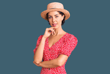 Young beautiful woman wearing summer hat looking confident at the camera with smile with crossed arms and hand raised on chin. thinking positive.