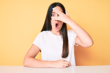 Young caucasian woman sitting at the desk wearing casual white tshirt peeking in shock covering face and eyes with hand, looking through fingers afraid