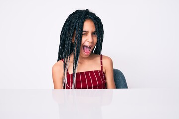 Young african american girl child with braids wearing casual clothes sitting on the table winking...