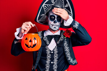 Young man wearing mexican day of the dead costume holding pumpkin stressed and frustrated with hand on head, surprised and angry face