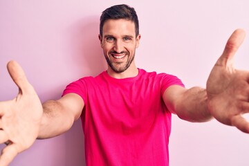 Young handsome man wearing casual t-shirt standing over isolated pink background looking at the camera smiling with open arms for hug. Cheerful expression embracing happiness.