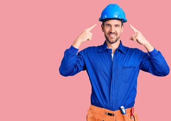 Young handsome man wearing worker uniform and hardhat smiling pointing to head with both hands finger, great idea or thought, good memory