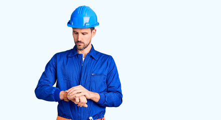 Young handsome man wearing worker uniform and hardhat in hurry pointing to watch time, impatience,...