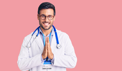 Young hispanic man wearing doctor uniform and stethoscope praying with hands together asking for...