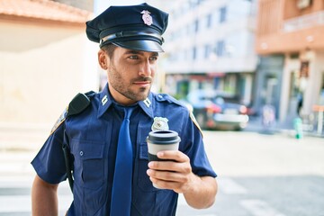 Young hispanic policeman wearing police uniform with serious expression. Drinking cup of take away coffee standing at town street.