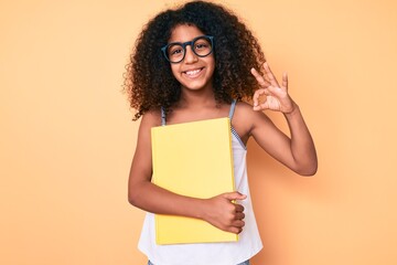 African american child with curly hair wearing glasses and holding book doing ok sign with fingers, smiling friendly gesturing excellent symbol