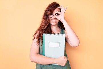 Young beautiful woman wearing glasses holding book smiling happy doing ok sign with hand on eye looking through fingers