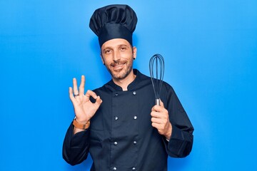 Young handsome man wearing cooker uniform holding whisk doing ok sign with fingers, smiling friendly gesturing excellent symbol