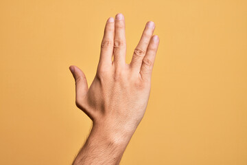 Hand of caucasian young man showing fingers over isolated yellow background greeting doing Vulcan salute, showing back of the hand and fingers, freak culture