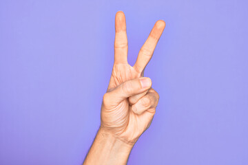 Hand of caucasian young man showing fingers over isolated purple background counting number 2 showing two fingers, gesturing victory and winner symbol
