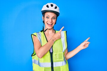 Young caucasian girl wearing bike helmet and reflective vest smiling and looking at the camera pointing with two hands and fingers to the side.