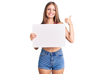 Fototapeta premium Young beautiful blonde woman holding blank empty banner smiling happy and positive, thumb up doing excellent and approval sign