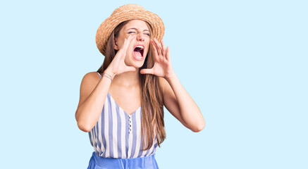 Young beautiful blonde woman wearing summer hat shouting angry out loud with hands over mouth